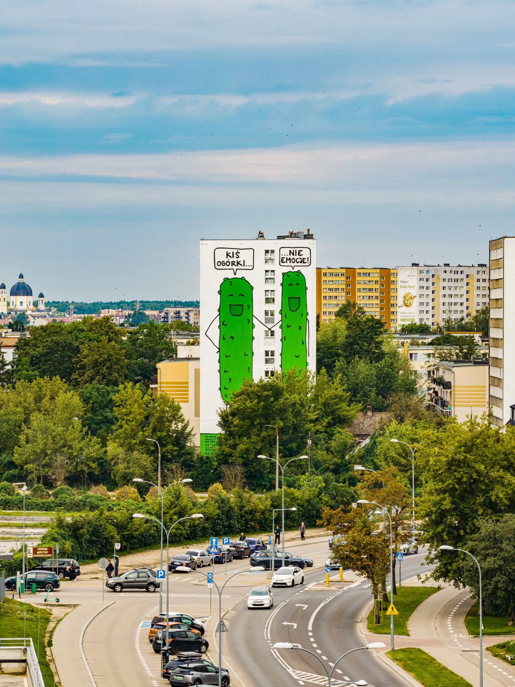 A white dormitory tower in Białystok with a mural of two green pickles and speech bubbles reading “Kiś ogórki…” and “…nie emocje!”, a street and trees in the foreground