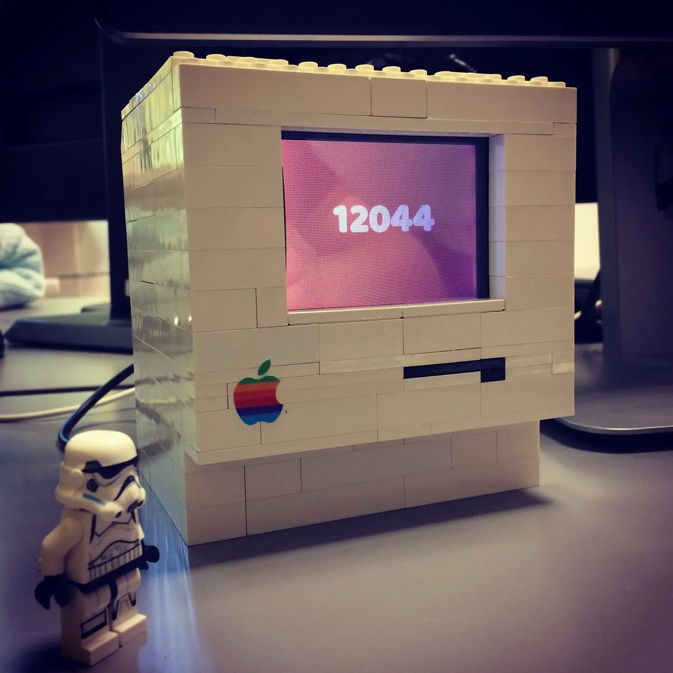 A white Macintosh 128K built from LEGO bricks with the rainbow Apple logo; the screen shows the number 12044, with a stormtrooper minifigure standing beside it