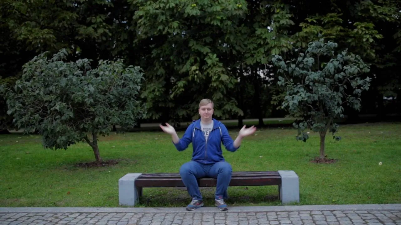 Film still: a man in a blue hoodie sits on a park bench, arms spread wide