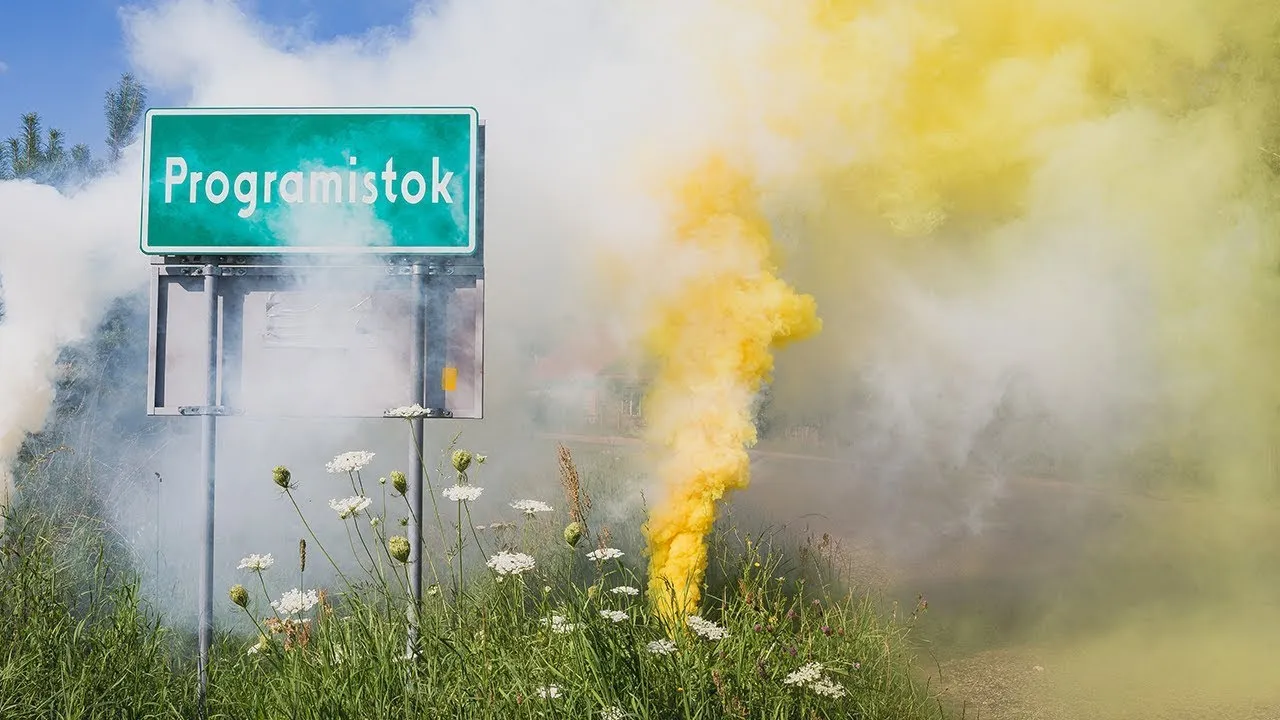 Green village sign reading “Programistok” engulfed in yellow and white flare smoke, in a meadow full of chamomile