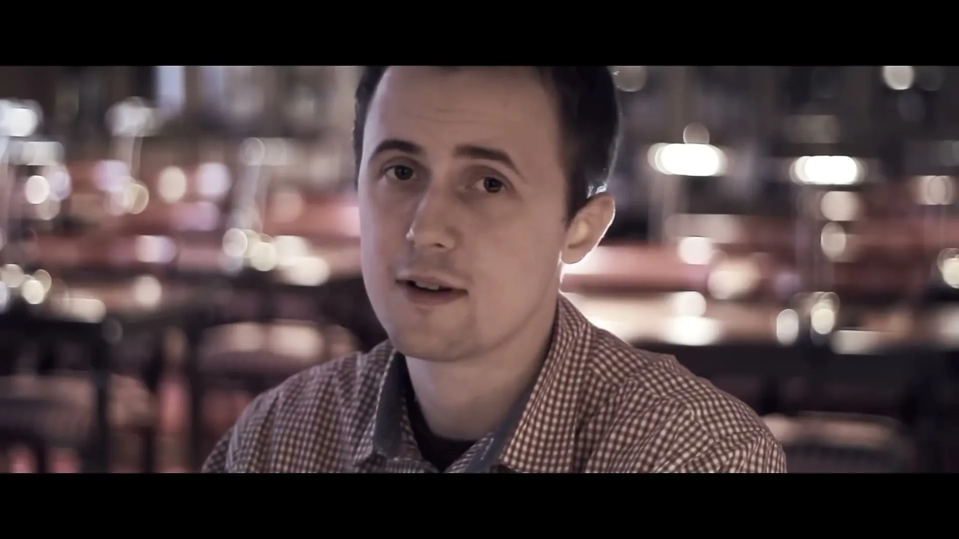 Still from the video: Dziemian in a dimly lit reading room, lamp lights blurred behind him