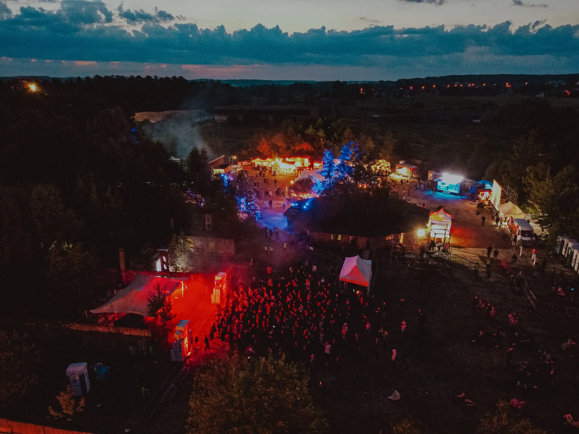 Night drone view of Up To Date Festival 2023: a crowd in red light in front of the stage, lit-up tents and forest all around